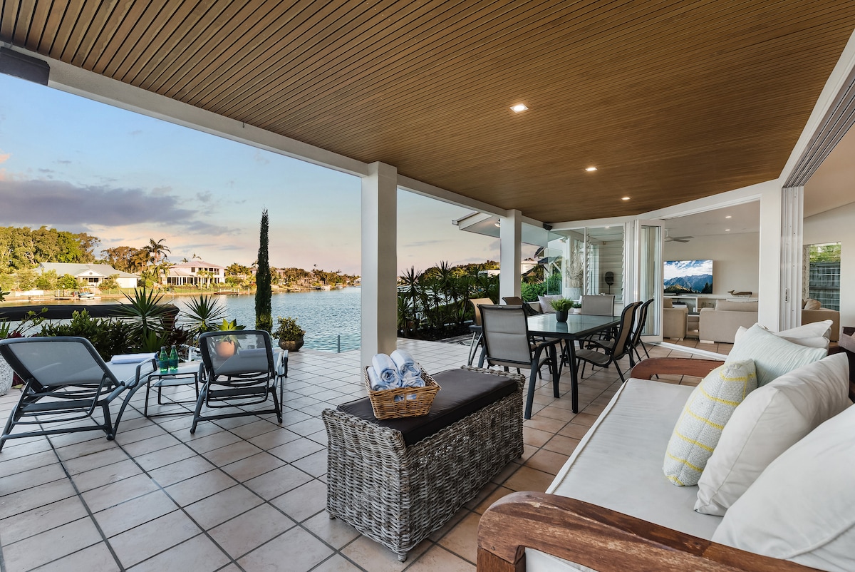 A spacious alfresco area features various seating options, including lounge chairs and a dining table. Potted plants add greenery, while views of the tranquil canal and waterfront are visible in the background. Soft light illuminates the area, creating a relaxing environment.