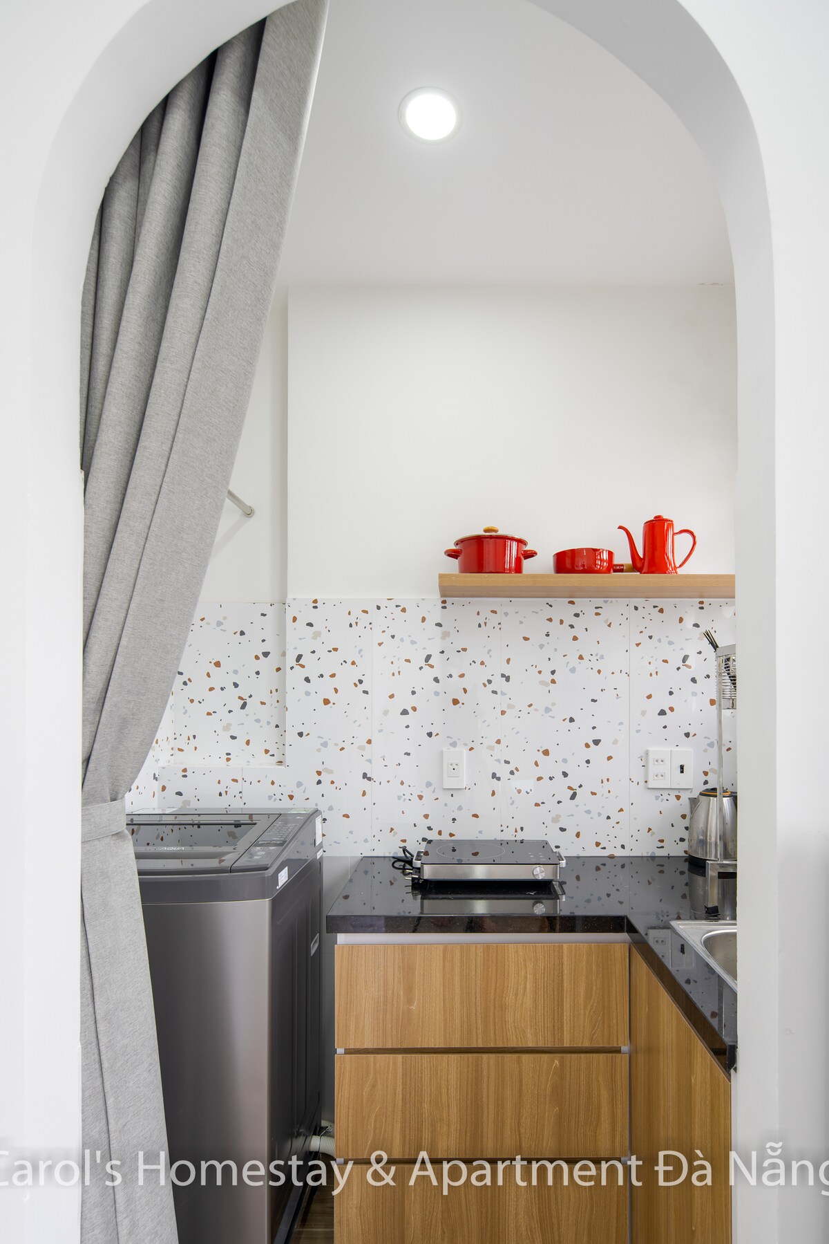 A modern kitchen area is presented, featuring a washing machine, a countertop with cooking utensils, and a bright backsplash adorned with colorful specks. Red kitchenware adds a pop of color, while warm wood cabinetry offers a sleek finish. Soft gray curtains provide a cozy touch.