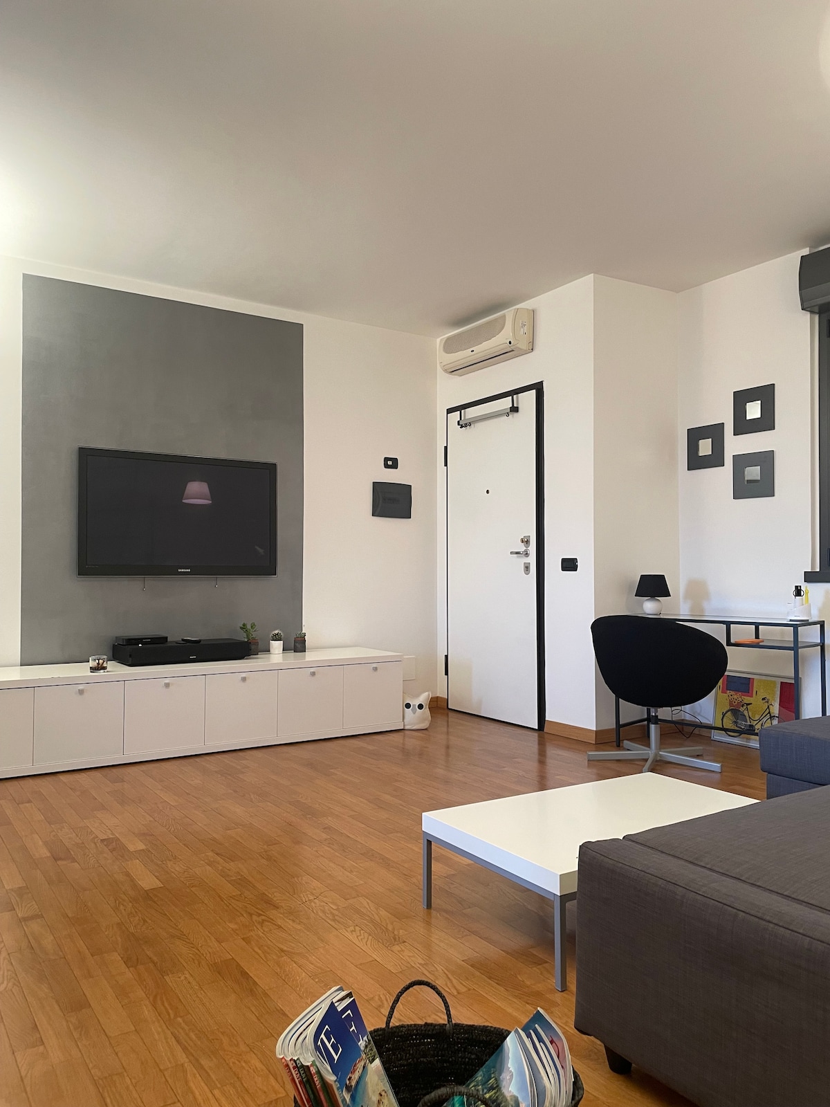 A living area features a mounted television above a low cabinet with a minimalist design. A black chair and a desk are positioned beside the wall, while a coffee table sits in the center. Natural light fills the space, enhancing the wooden flooring.