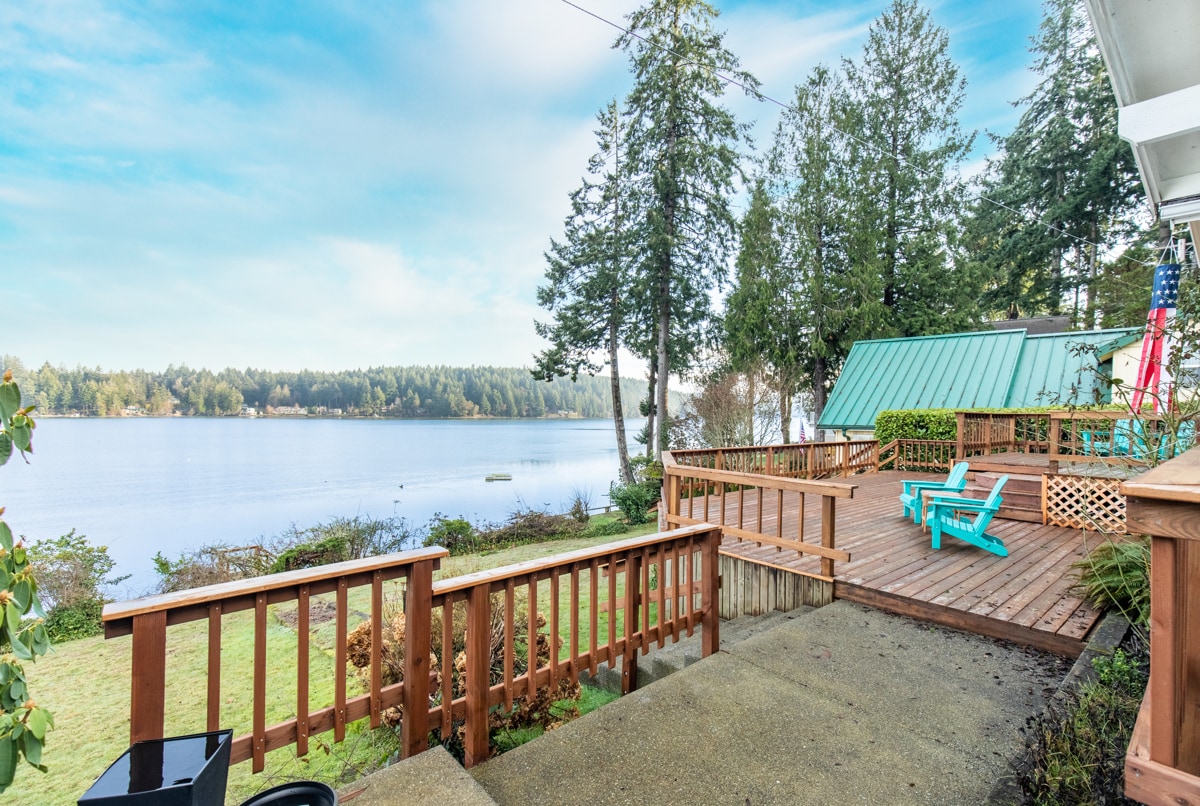 A serene view of the bay is visible from the expansive wooden deck, featuring vibrant blue chairs. Lush green trees frame the scene, enhancing the tranquil ambiance. A gently sloped yard leads down to the water, inviting moments of relaxation and enjoyment.