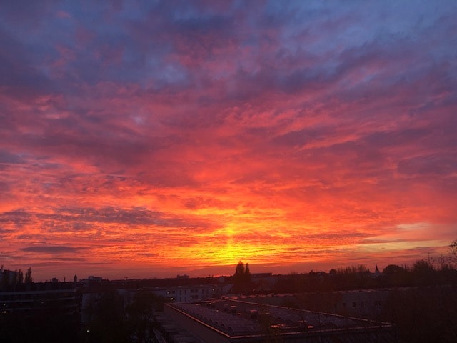 A vibrant sunset is displayed, with a palette of oranges, pinks, and purples filling the sky. The sun is positioned near the horizon, casting a warm glow over the landscape below. The image captures the expansive view from the top floor, emphasizing the serene evening atmosphere.