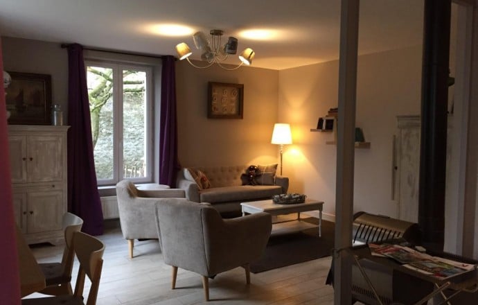 A comfortable living area features a beige sectional sofa and a pair of armchairs, arranged around a coffee table. Natural light enters through large windows, while a soft light fixture adds warmth. A decorative cabinet and wall shelves provide additional charm.