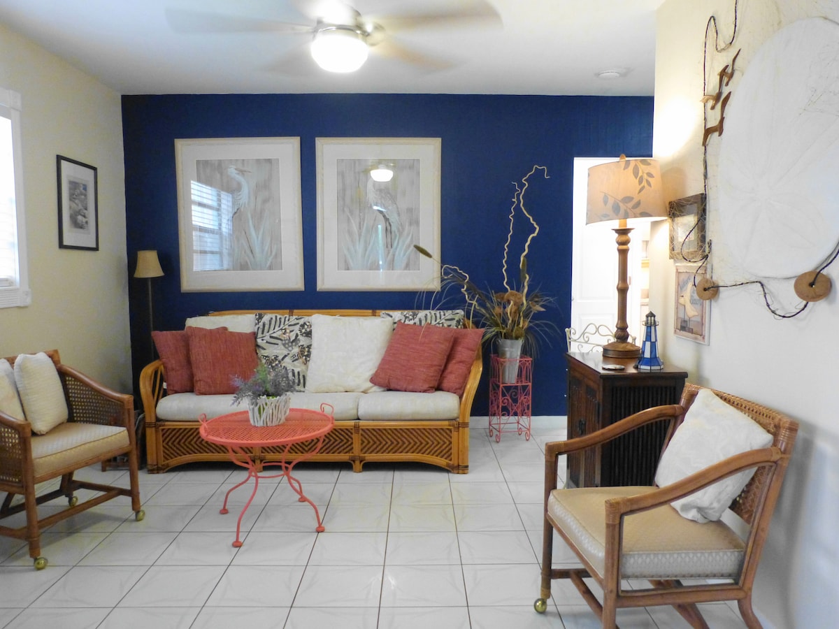 The living area features a comfortable seating arrangement with a sofa adorned with pillows, complemented by two wicker chairs. A round coffee table sits at the center, and artwork is displayed on the wall in muted tones, enhancing the room's relaxed ambiance.