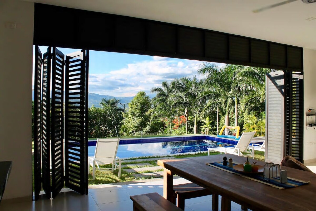 Large glass doors open to a serene outdoor space with a pool surrounded by lush greenery. Two lounge chairs are positioned beside the water. A wooden dining table is visible in the foreground, with a set of colorful tableware arranged neatly.