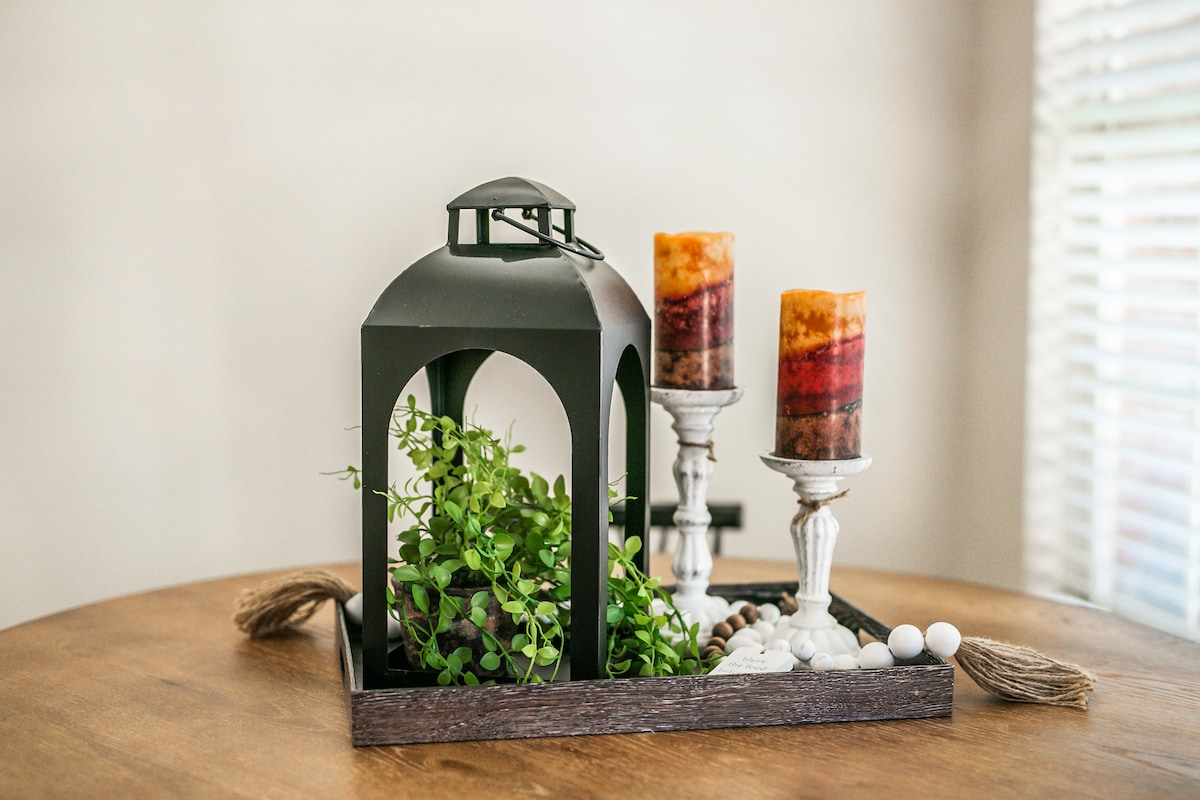 A decorative centerpiece is presented on a wooden table, featuring a lantern encasing green foliage. Two candles in amber tones are mounted on white candle holders, complemented by a tray of smooth stones and decorative accents, creating a serene setting.