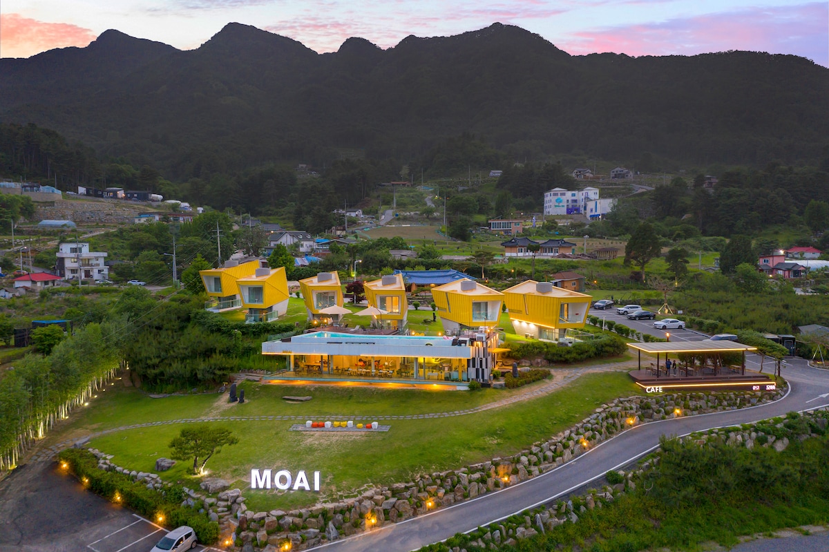 The exterior view showcases a cluster of modern yellow structures set against a backdrop of rolling mountains. Lush greenery surrounds the building, with lighting accentuating pathways and the MOAI sign. A well-kept landscape contributes to the inviting appearance of the property.