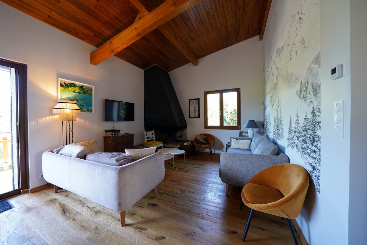 A spacious living area features a large window allowing natural light to fill the room. A modern fireplace stands against one wall, complemented by comfortable seating options, including a cozy sofa and distinctive armchairs. Wooden beams and flooring enhance the warm ambiance.