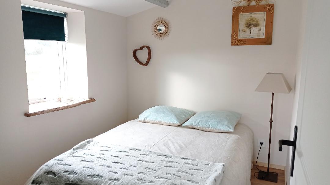 A serene bedroom features a spacious bed with light blue pillows. Natural light enters through a window adorned with a dark shade. Decorative elements include a round mirror, a heart-shaped wall hanging, and a framed artwork above the bed, alongside a bedside lamp.