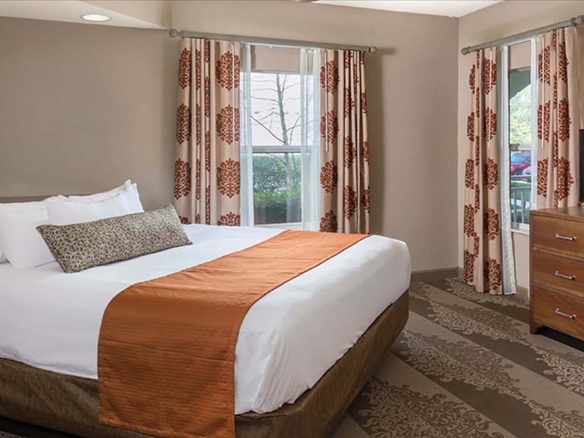 A well-lit bedroom features a spacious bed dressed in crisp white linens and an orange bed runner. Large windows allow natural light to fill the room, and patterned curtains frame the view. A wooden nightstand is positioned next to the bed, adding warmth to the space.