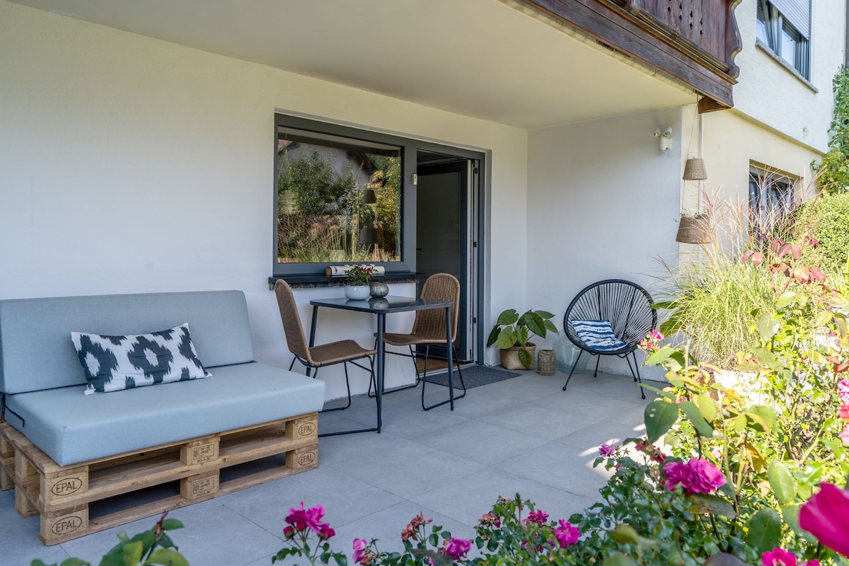 A furnished terrace provides a relaxing outdoor space, featuring a cozy seating area with a pallet couch and a small dining table. Lush plants and vibrant flowers adorn the area, creating a welcoming atmosphere near the entrance to the accommodation.