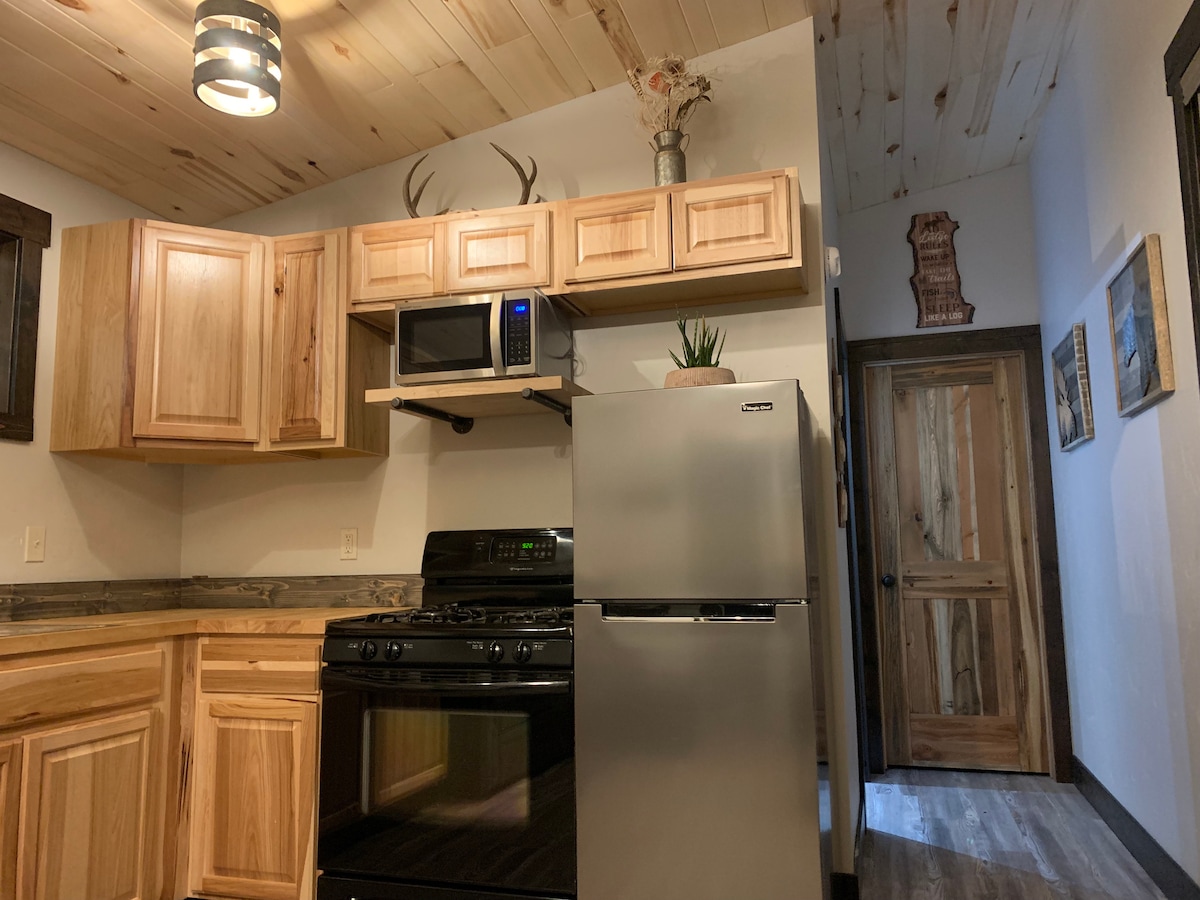 A functional kitchen is showcased, featuring wooden cabinets and modern appliances. A gas stove, microwave, and stainless steel refrigerator are visible, with a small decorative plant on the countertop. The door and walls reveal natural wood elements, contributing to a warm, inviting aesthetic.