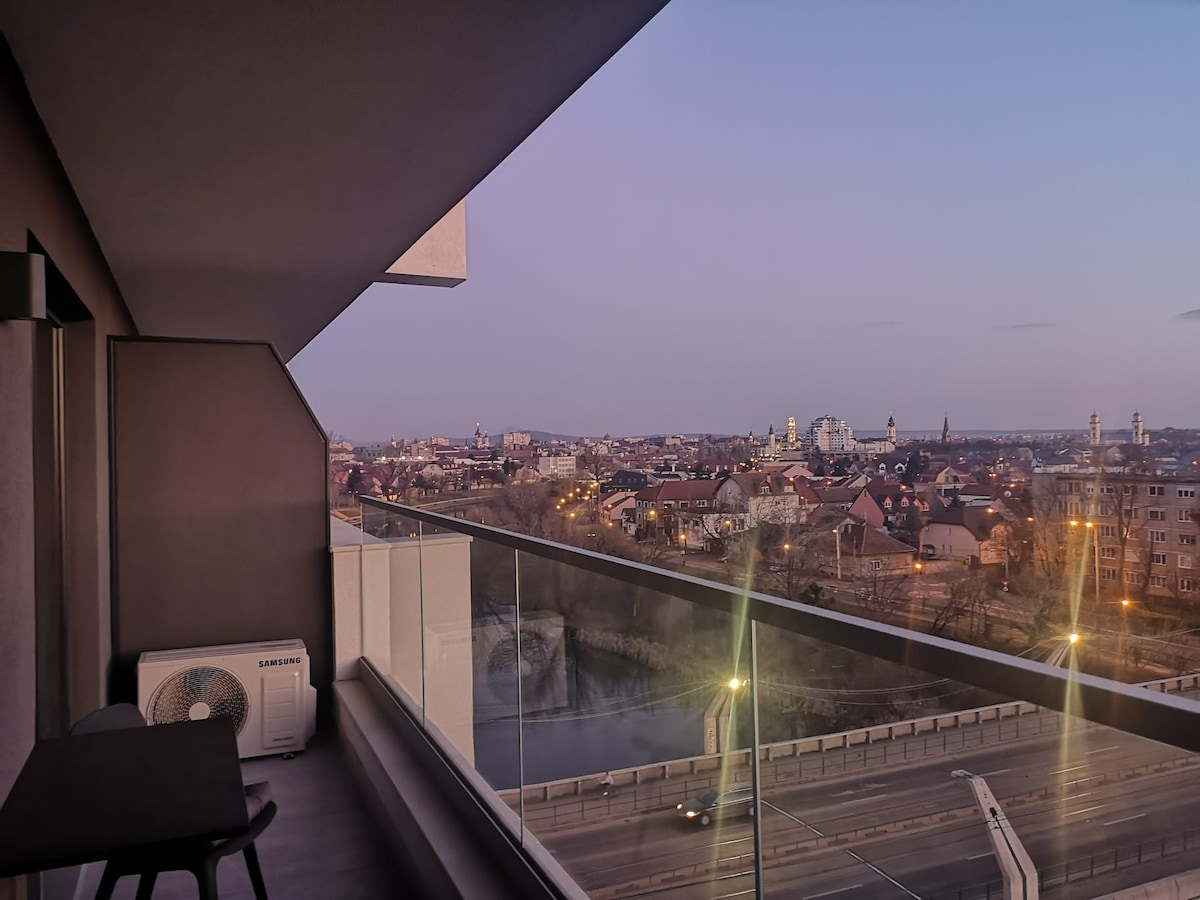 A balcony is shown with a sleek design overlooking the cityscape. Glass railings provide an unobstructed view of the buildings and streets below, capturing the essence of urban life as the evening light casts a soft glow over the scene.