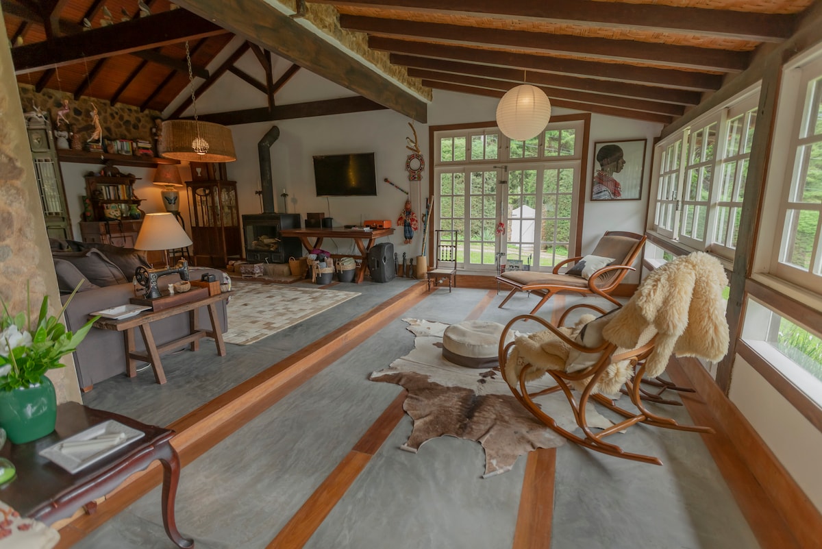 A cozy living area features exposed wooden beams and large windows, allowing natural light to fill the space. Comfortable seating, including a rocking chair and plush sofa, is arranged on a mix of textures, such as a soft area rug and hardwood flooring.
