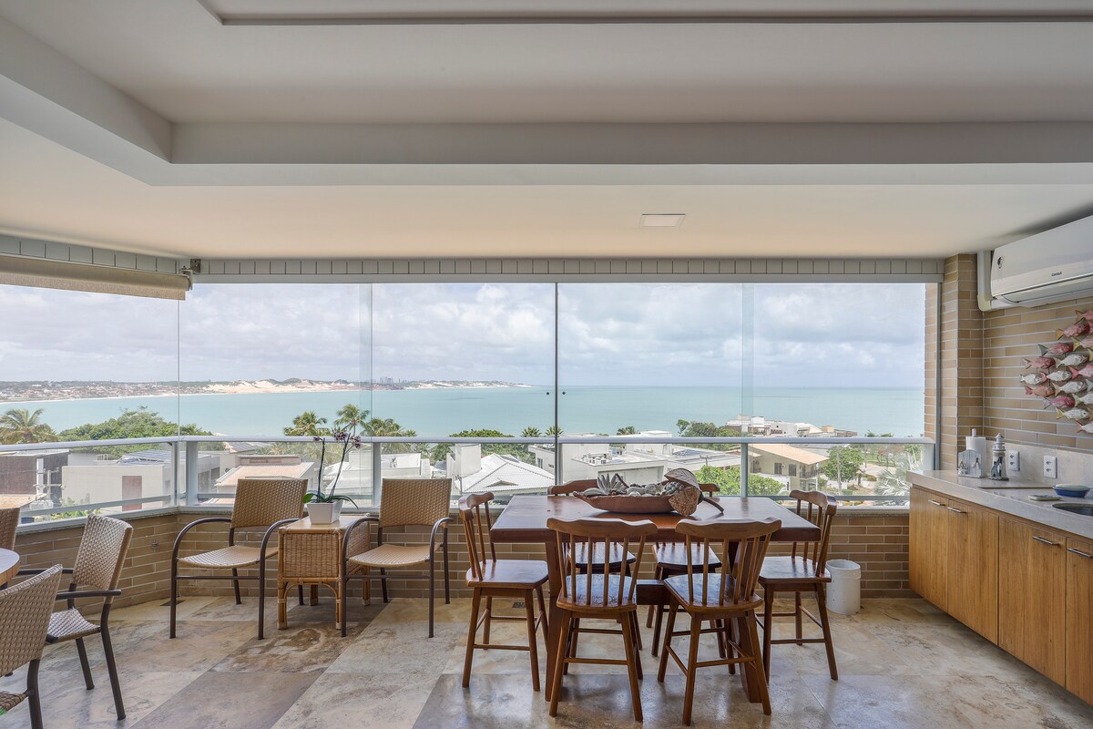 A spacious dining area is equipped with wooden tables and chairs, overlooking a panoramic view of the ocean and sandy dunes through large glass windows. Natural light fills the room, creating an inviting atmosphere.