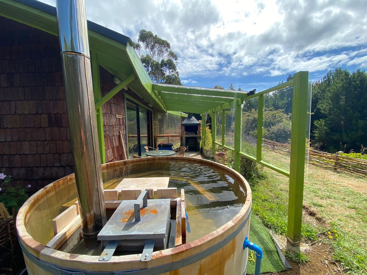 A wooden hot tub is featured on a green terrace, complimented by a metal chimney. Natural surroundings include trees and open fields, with a partially covered area providing shade. Clear skies and soft clouds create a serene backdrop for relaxation.