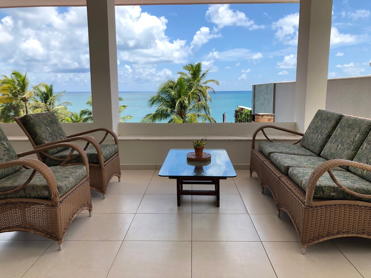 A seating area is presented with two sets of rattan sofas and a coffee table, overlooking a serene ocean view. Green palm trees are visible in the foreground, while light cloud formations add depth to the clear blue sky.