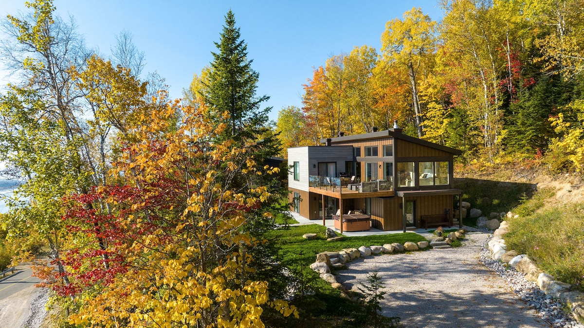 A modern chalet is surrounded by vibrant autumn foliage, featuring large windows that showcase convenient outdoor access. The well-maintained landscape includes a gravel pathway and natural stone accents, while a hot tub is visible on the terrace, inviting relaxation amidst nature.