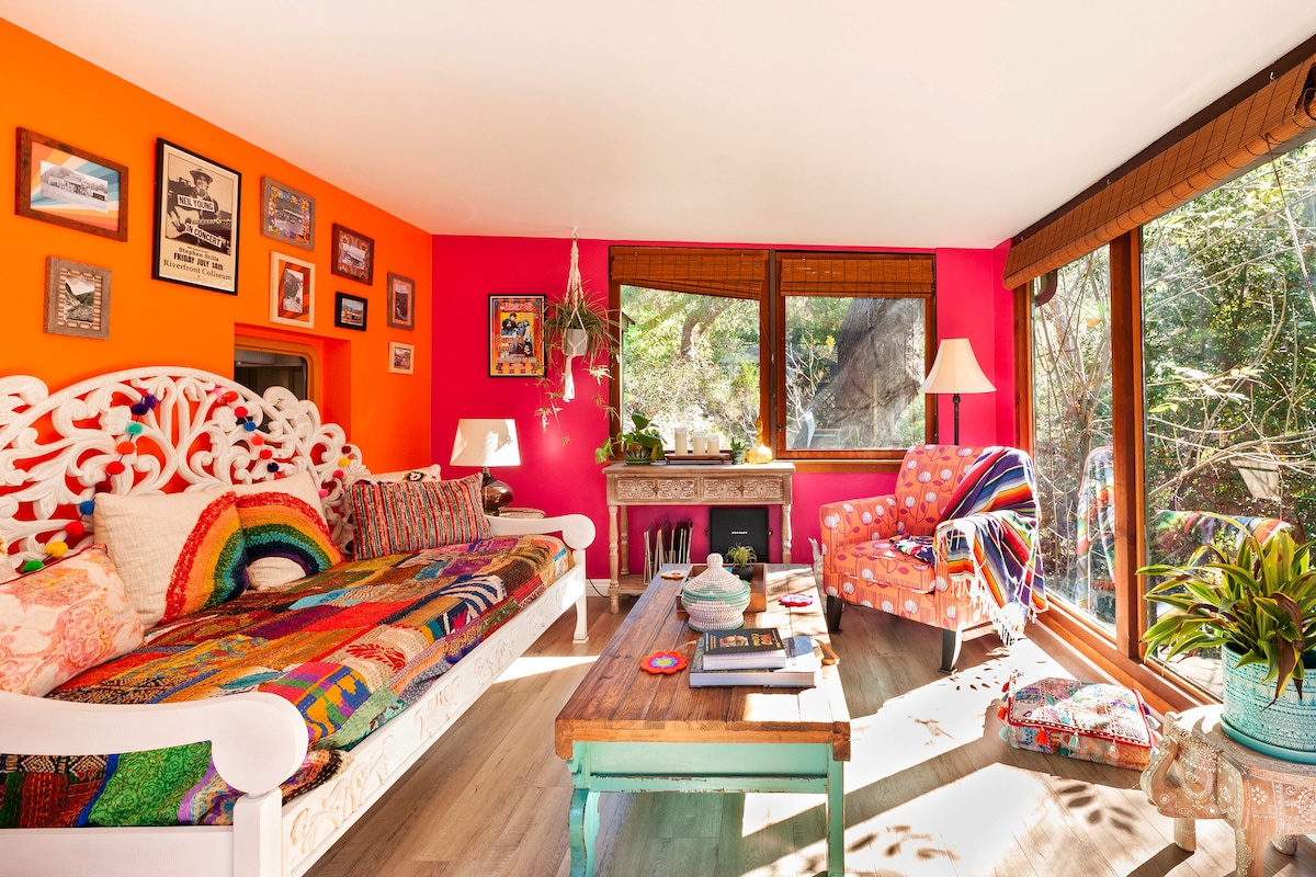 A bright and lively great room features walls in vibrant orange and pink. A colorful sofa adorned with a rainbow blanket invites relaxation, complemented by an armchair in a coordinating fabric. Large windows with natural light overlook surrounding greenery, creating a cheerful and cozy atmosphere.