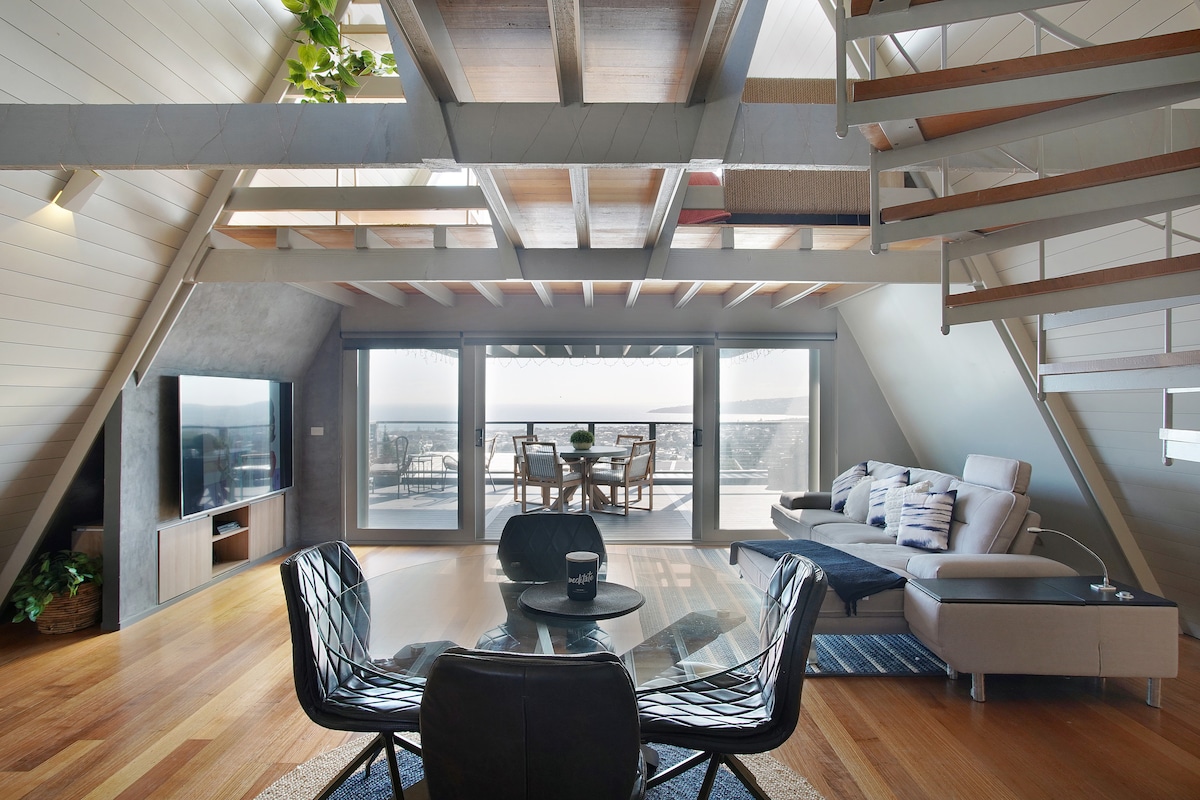 An open-plan living space features a round dining table surrounded by stylish black chairs. A comfortable sectional sofa offers a relaxed seating area, with large windows allowing natural light and ocean views. The unique A-frame architecture highlights wooden beams and a staircase leading to the upper level.
