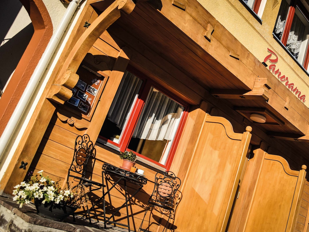 A wooden exterior features an inviting entrance adorned with decorative carvings. A small bistro set, consisting of two wrought-iron chairs and a round table, is situated on the covered porch. A potted plant adds a touch of greenery to the scene, framed by flower boxes.