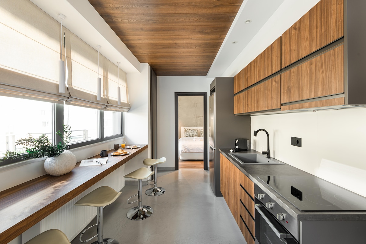 A modern kitchen area showcases wooden cabinetry and sleek black countertops, complemented by barstools and a dining nook by the window. Natural light fills the space, with a view leading to a cozy bedroom in the background, enhancing the open layout.
