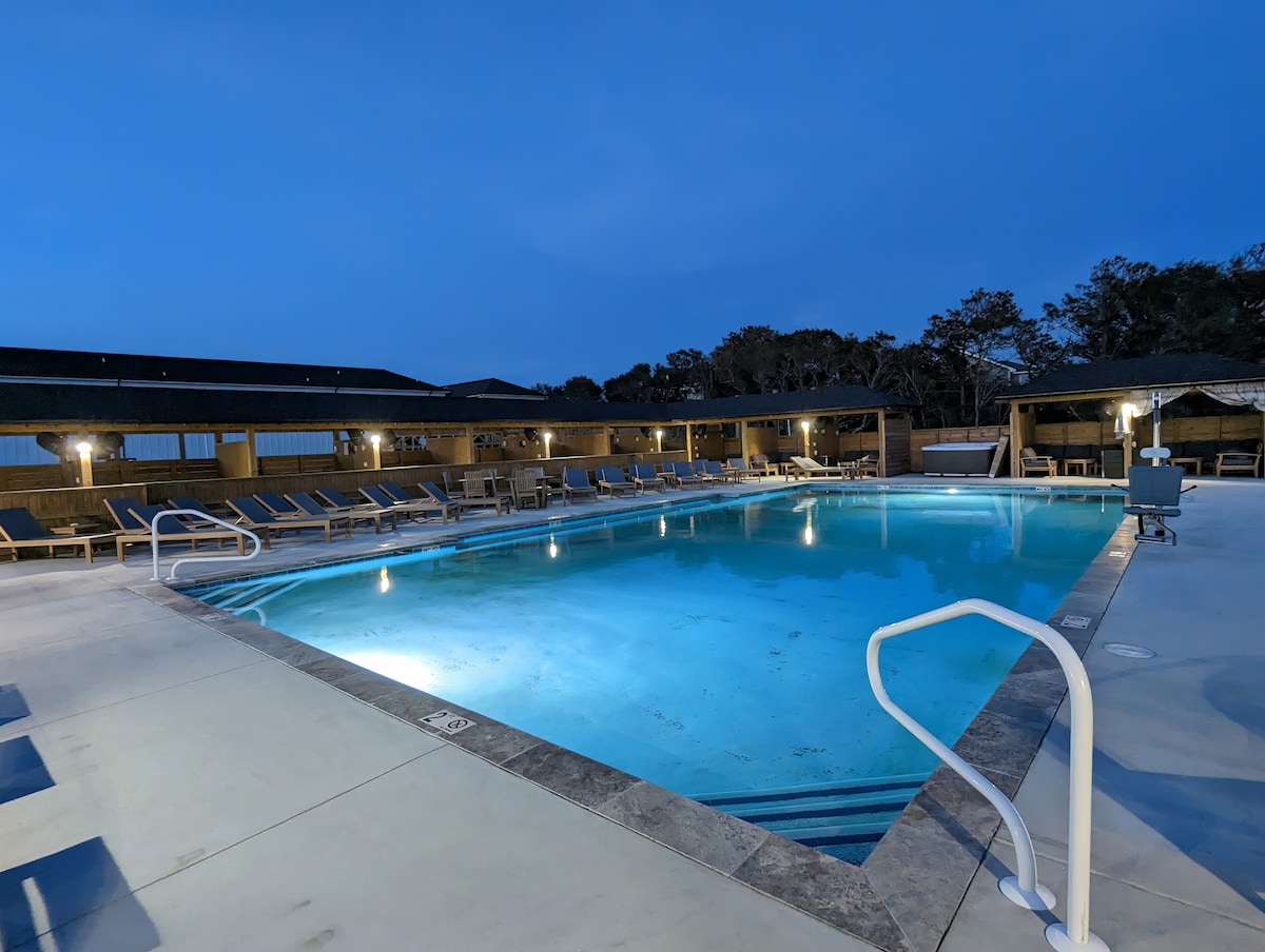 A well-lit, heated community swimming pool is presented, surrounded by lounge chairs. The pool is complemented by a spacious deck area and is bordered by a wooden fence, creating a private ambiance. The evening sky reflects softly on the water's surface.