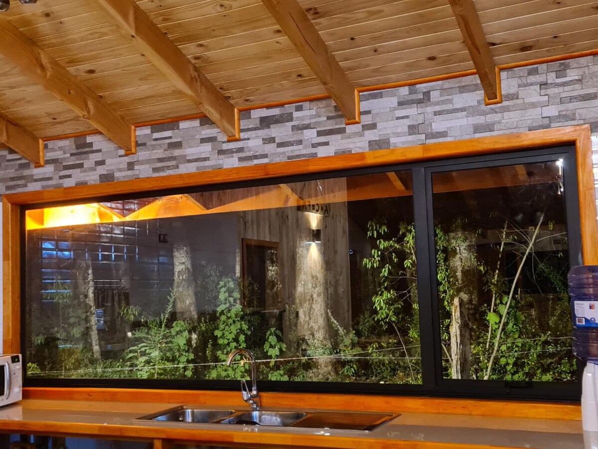 A large window frames a view of lush greenery outside, allowing natural light to fill the kitchen space. The wooden beams of the ceiling complement the stone-accented walls, creating a warm and inviting environment. The sink is positioned under the window for a tranquil cooking experience.