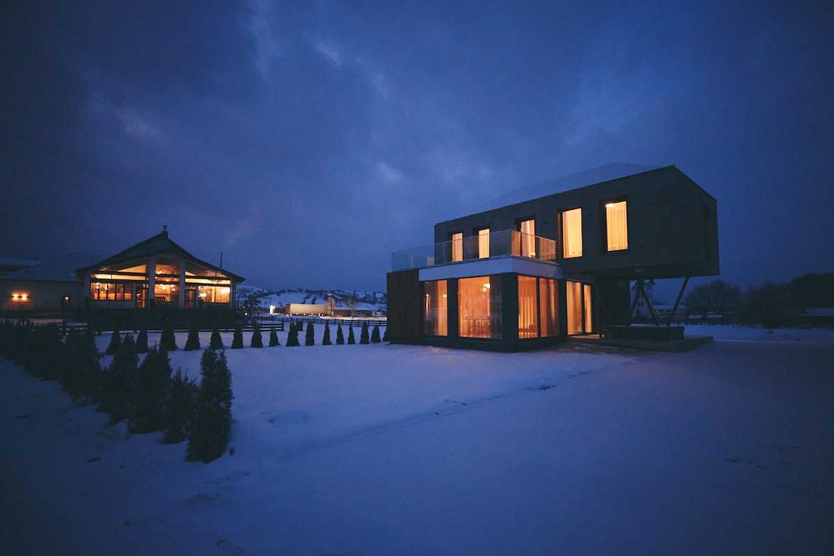 A modern, two-story building is showcased against a twilight sky, with soft lighting emanating from expansive glass windows. The surrounding landscape is covered in snow, while a neighboring structure features a traditional architectural style, adding to the visual contrast.
