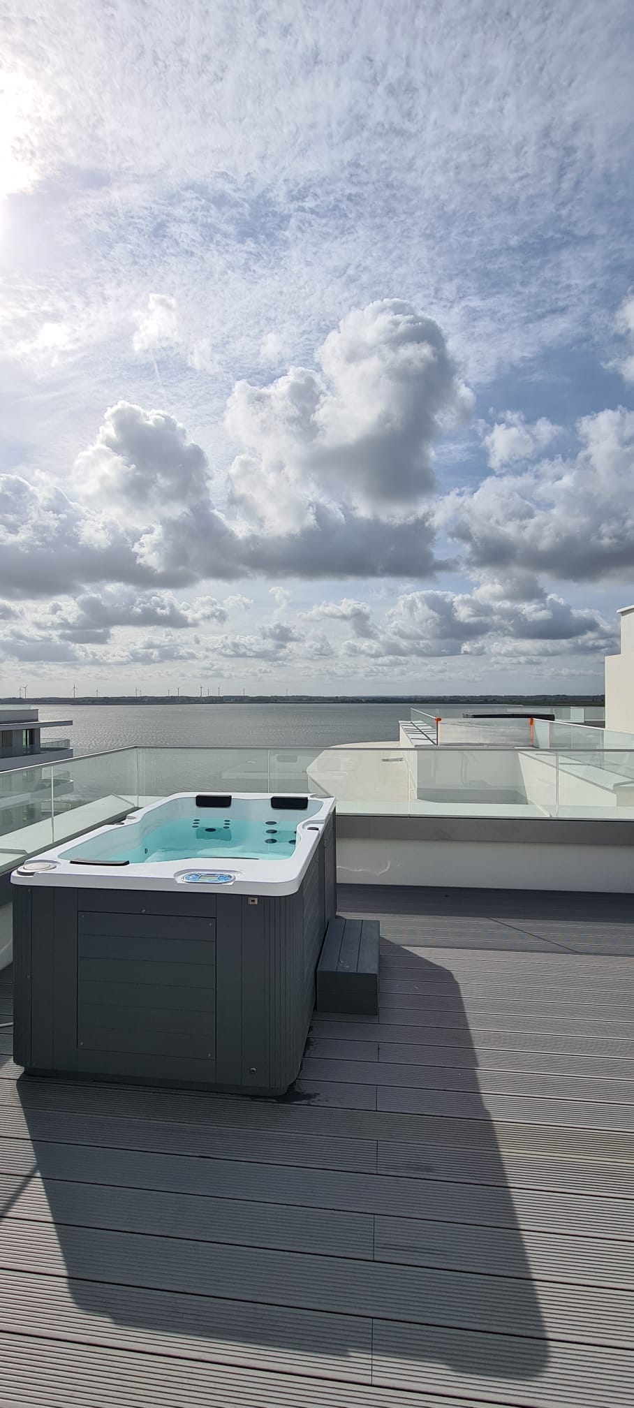 A rooftop terrace features a Jacuzzi surrounded by wooden decking, offering views of the expansive sky and serene water. The space is designed for relaxation, with clear glass barriers enhancing the open atmosphere.