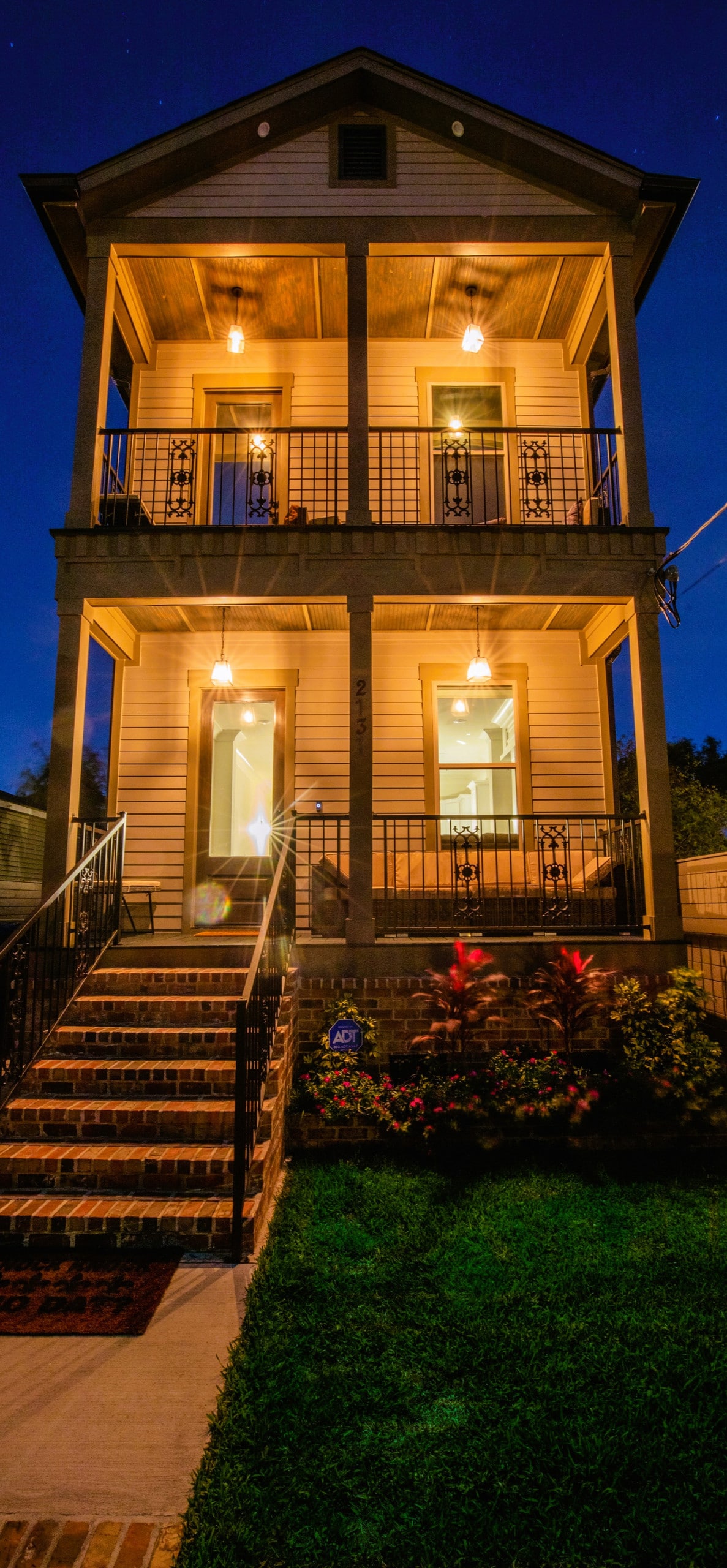 A two-story home is showcased in evening light, featuring symmetrical windows and a welcoming entry. Decorative outdoor lighting highlights the staircase leading to the porch, which offers a view of the well-maintained front yard with vibrant plants.