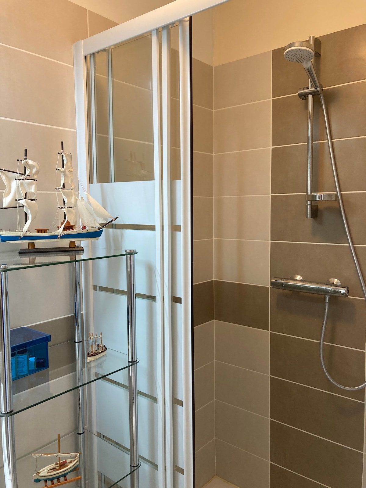 A modern shower area is enclosed with glass doors and features a handheld shower head. Shelves nearby display decorative miniature sailboats and a blue container. The tiled walls are in soft neutral tones, enhancing the clean and contemporary feel of the space.