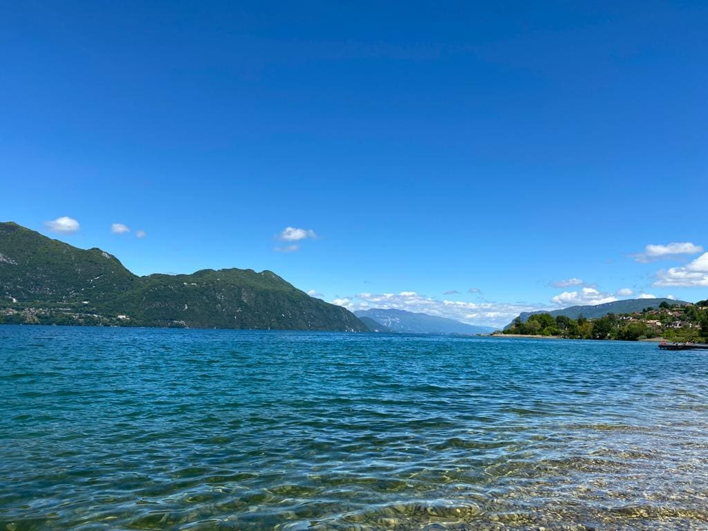 A clear blue lake spans the image, reflecting the bright sky filled with scattered clouds. Green hills frame the water's edge, leading to gently sloping mountains in the distance. The shoreline features pebbles merging with soft sand, creating a natural gradient along the water.