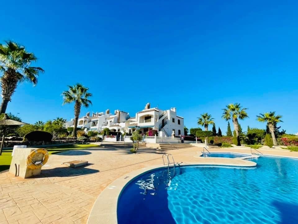 A view of the landscaped grounds surrounding the penthouse, featuring a large swimming pool with a gently sloping entrance. Palm trees and garden areas add greenery, while a white building with multiple stories stands in the background under a clear blue sky.