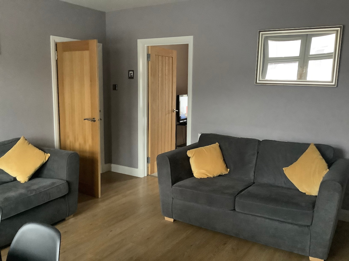 A living area is shown with two gray sofas, each featuring yellow accent pillows. Light hardwood flooring complements the soft gray walls. A rectangular mirror reflects natural light, and a wooden door partially ajar leads to another room.