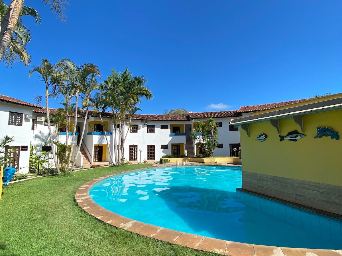 A curved swimming pool is surrounded by a lush green lawn and inviting palm trees. The white and yellow buildings form a circular layout in the background, offering a serene atmosphere under a clear blue sky.