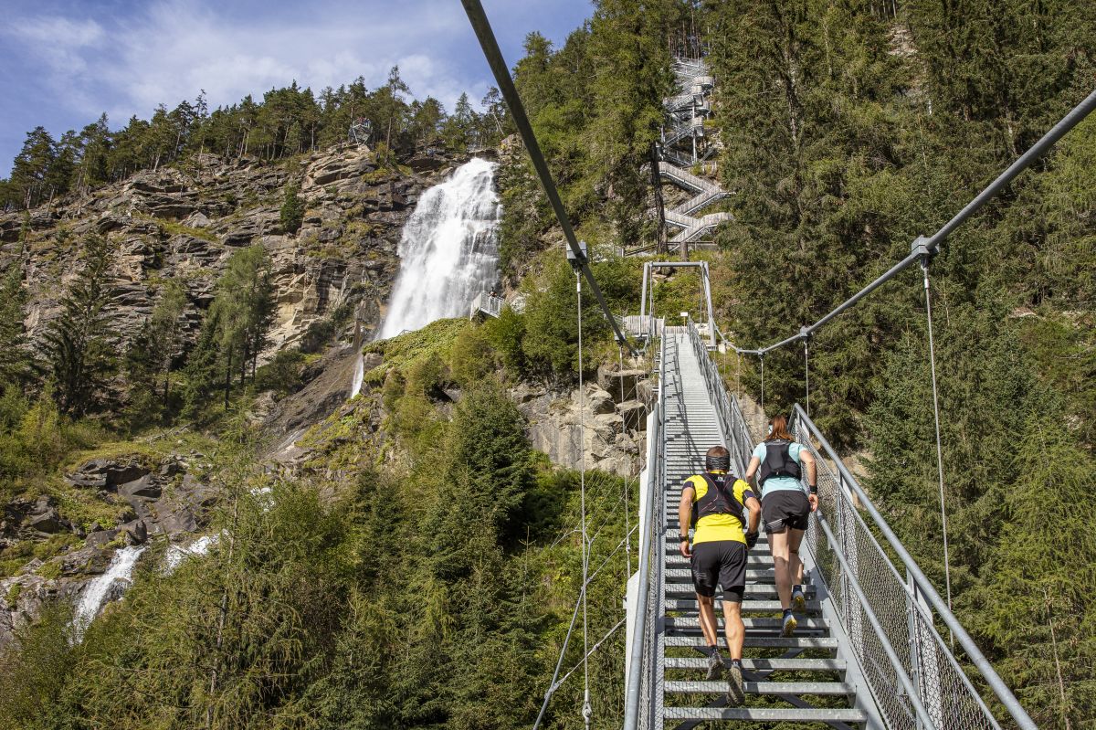 A suspension bridge is seen, leading upwards through a lush green landscape, with a waterfall cascading in the background. Two individuals are ascending the metal stairs, surrounded by tall trees and rocky terrain that enhances the natural scenery.