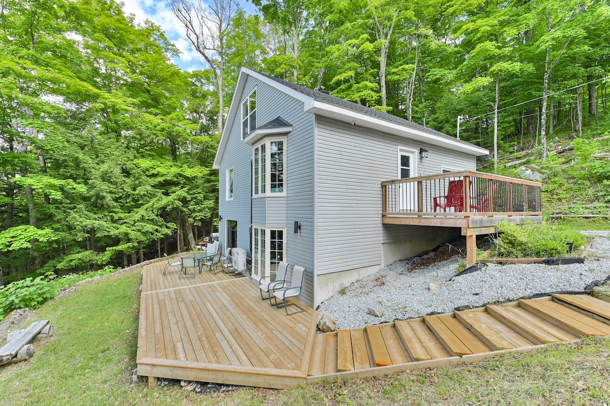The exterior of the cottage features a spacious wooden deck surrounded by lush greenery. A seating area with chairs is arranged on the deck, offering a relaxing outdoor space. Large windows enhance the natural light, creating a warm and inviting environment.