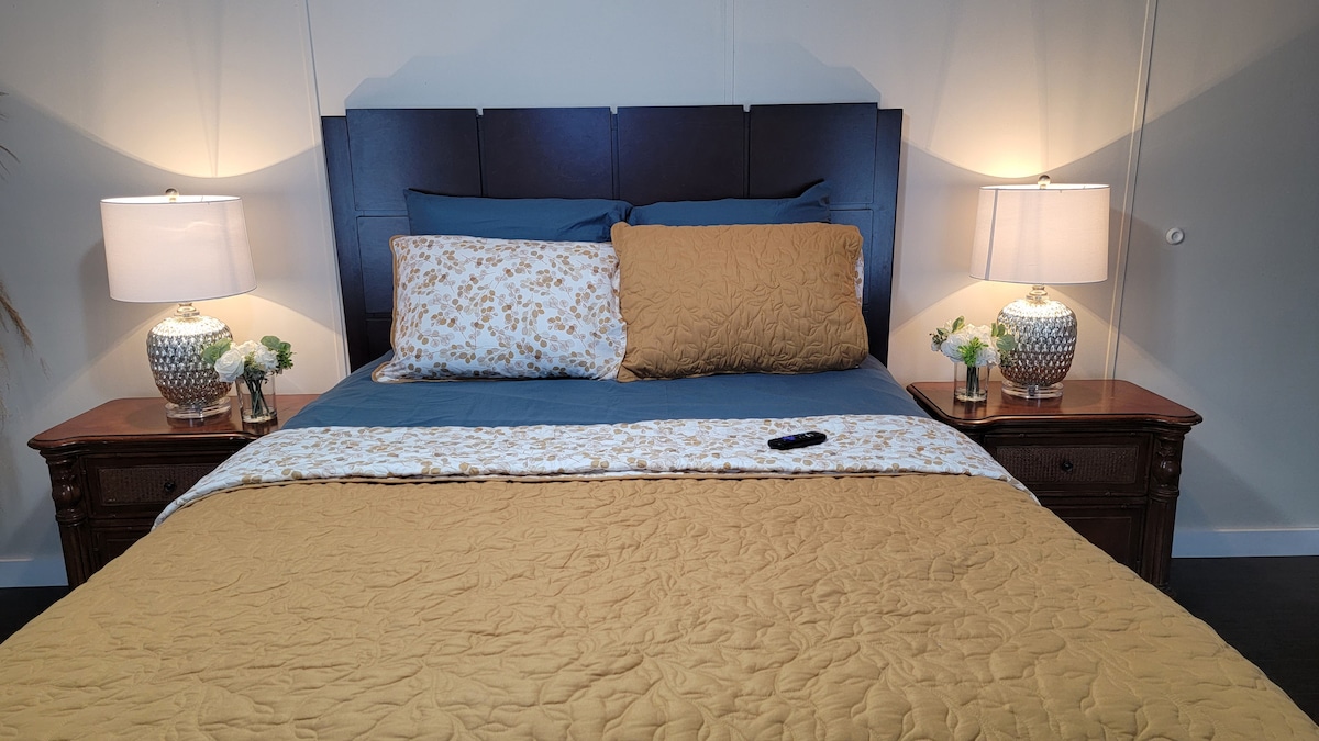 A spacious bed is centered against a wall, featuring a dark headboard and layered with an assortment of pillows. Nightstands with decorative lamps sit on either side, complemented by small vases of flowers. A remote control rests on the bedspread.
