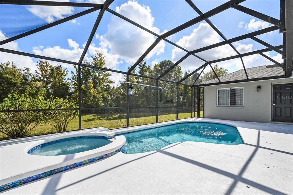 The outdoor area features a screened-in pool surrounded by a spacious deck. A jacuzzi is integrated into the pool area, providing a relaxing option. Lush greenery is visible beyond the enclosure, enhancing the outdoor experience.