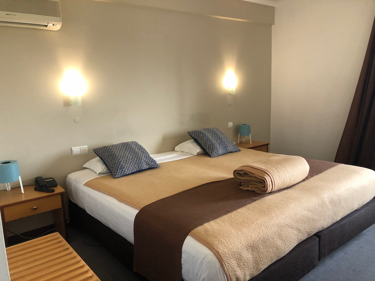 A spacious sleeping area features two beds dressed in light linens, complemented by decorative pillows. A warm-toned blanket is neatly rolled on top. Bedside tables with soft blue lamps are positioned on either side. A wall-mounted air conditioning unit is visible above the beds.