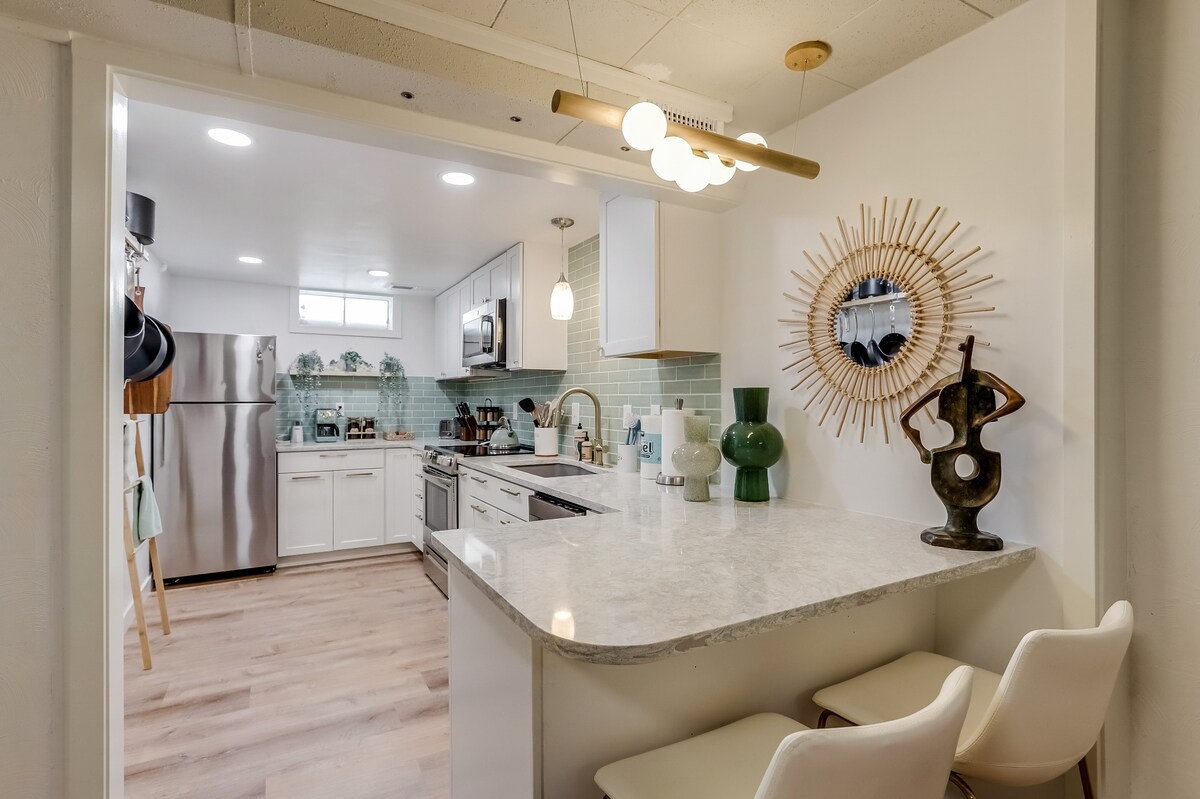 A modern kitchen features a spacious island with two high-backed stools, surrounded by stainless steel appliances and white cabinetry. Soft green tiles accent the back wall, while pendant lighting adds warmth to the inviting cooking area.