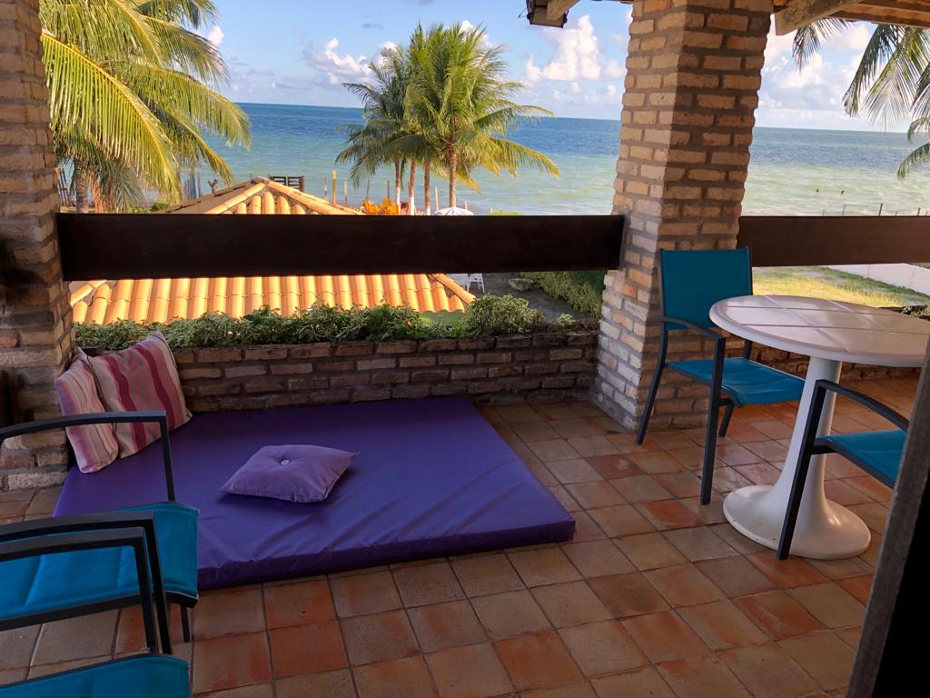 A shaded terrace features a purple mat for relaxation, accompanied by two blue chairs and a small round table. Lush greenery and vibrant coastal views are visible beyond the terrace, enhancing the inviting outdoor space.