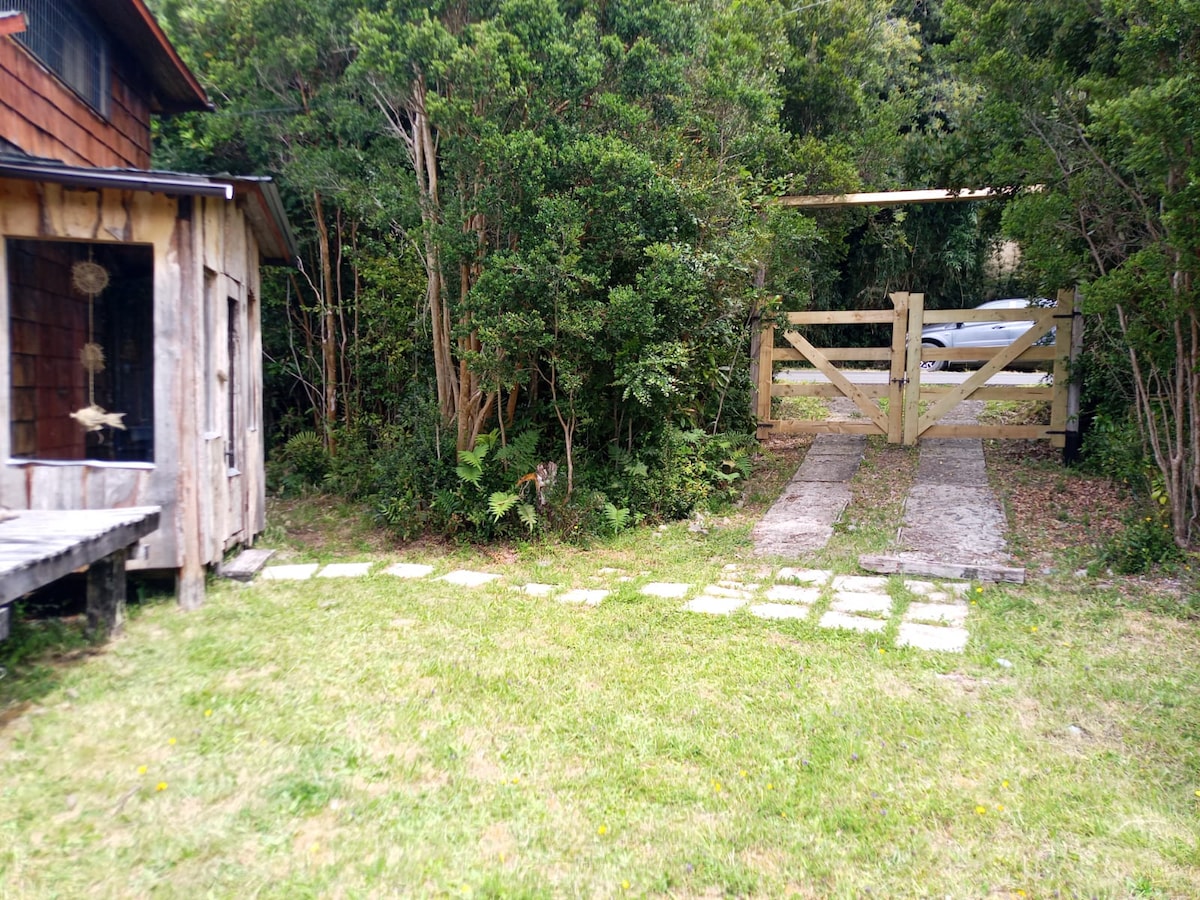 A pathway made of flat stones leads to a wooden gate, framed by dense greenery typical of the surrounding natural environment. A vehicle is parked just beyond the gate, offering easy access to the property. Grass is well-maintained in the area surrounding the path.