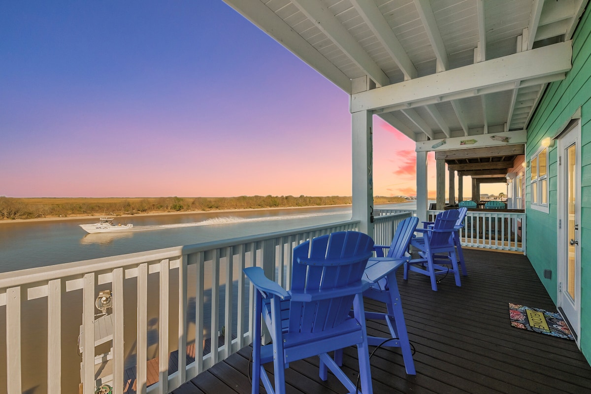 A spacious deck featuring two blue Adirondack chairs overlooks the Colorado River. The scene is enhanced by a vibrant sunset with shades of pink and orange in the sky. A boat glides smoothly across the water, completing the picturesque view.