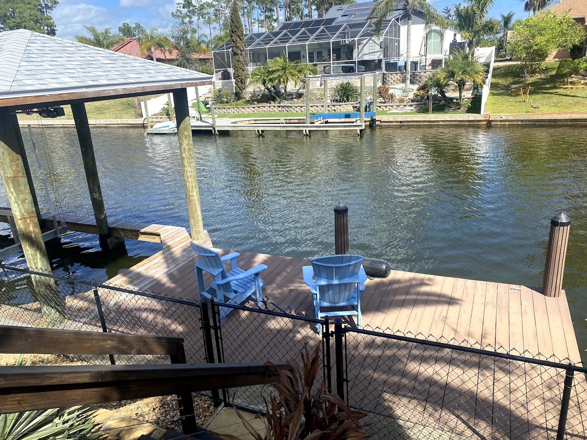 A wooden dock features two light blue chairs positioned beside the tranquil waterway. Lush greenery and a well-maintained property are visible across the canal, creating a peaceful setting for relaxation or fishing.