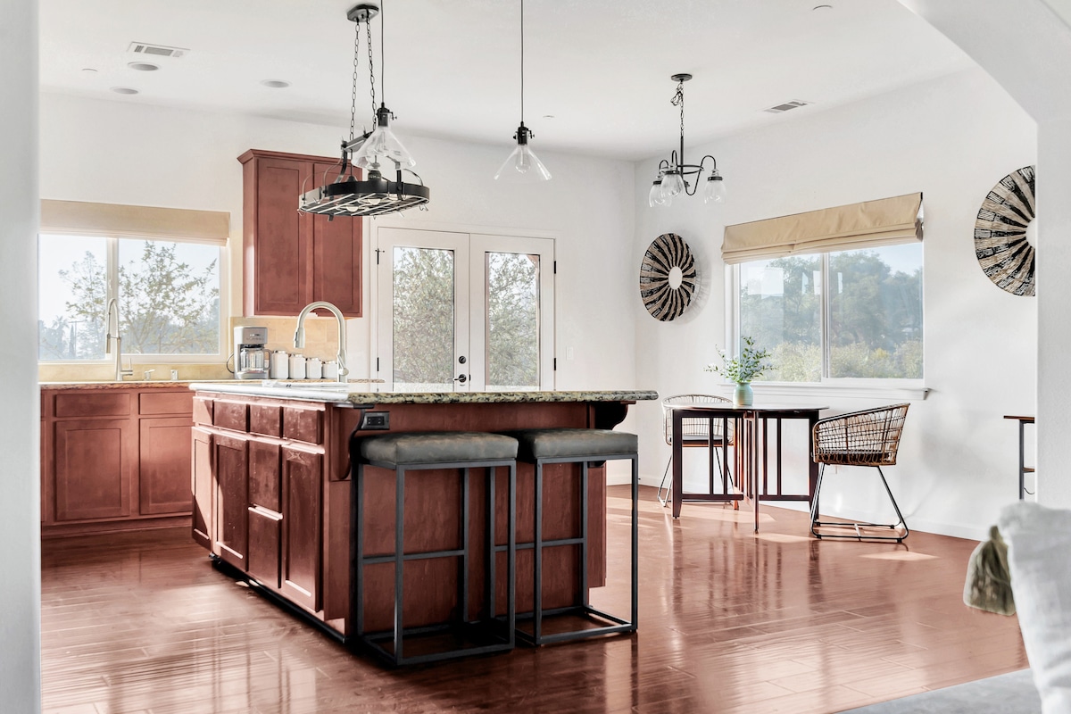 An open kitchen and dining area features a large island topped with granite, complemented by two metal stools. Natural light floods the space through multiple windows, illuminating the warm wood cabinetry. A small dining table and an accent chair create additional seating options.