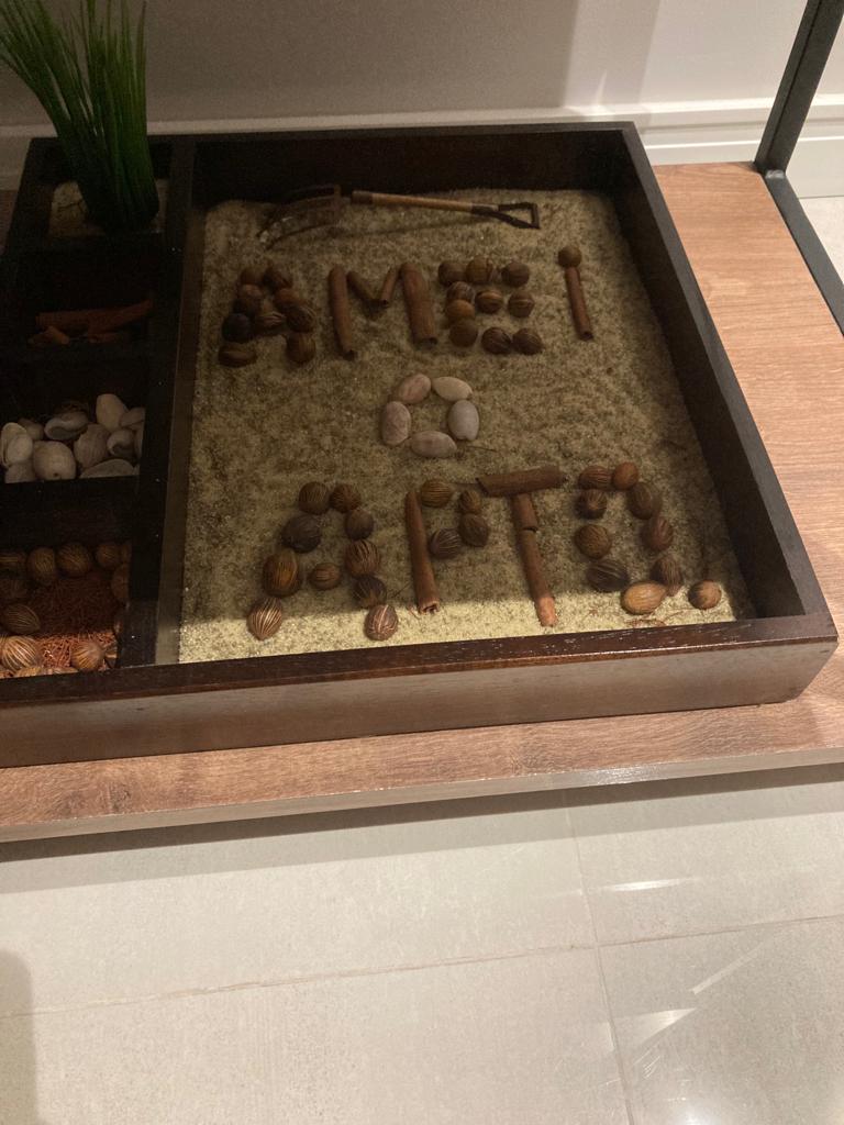 A decorative tray filled with sand features creatively arranged elements including pebbles, cinnamon sticks, and shells. The letters 'AMOR' are formed from pebbles, adding an artistic touch to the display.