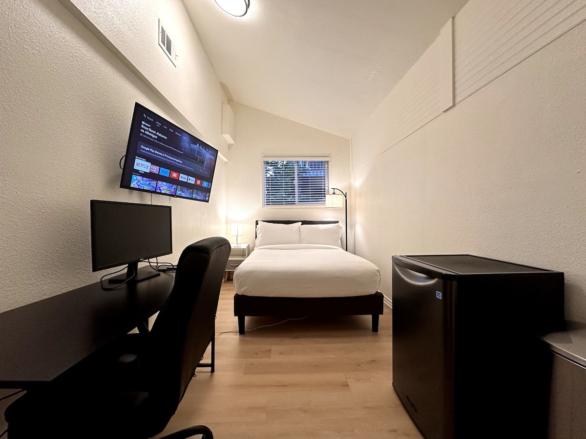 A private room features a spacious bed, a flat-screen TV mounted on the wall, and a work desk with an ergonomic chair. A mini-fridge stands in the corner. Natural light filters in through a window with partially closed blinds, complementing the neutral-colored walls.