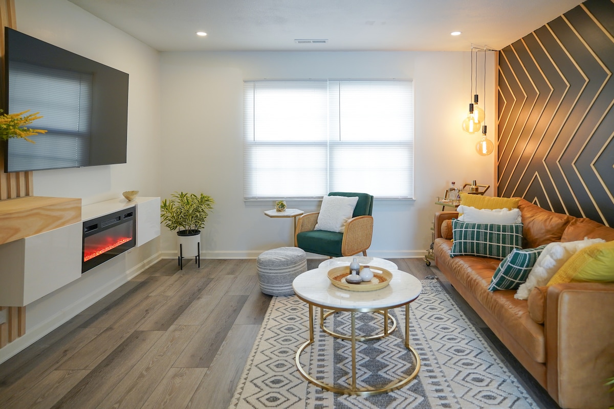 A welcoming living room features a leather sofa and a cozy armchair, complemented by a round coffee table. Natural light filters through the windows, while a geometric accent wall adds a modern touch. Decorative items and plants enhance the inviting atmosphere.