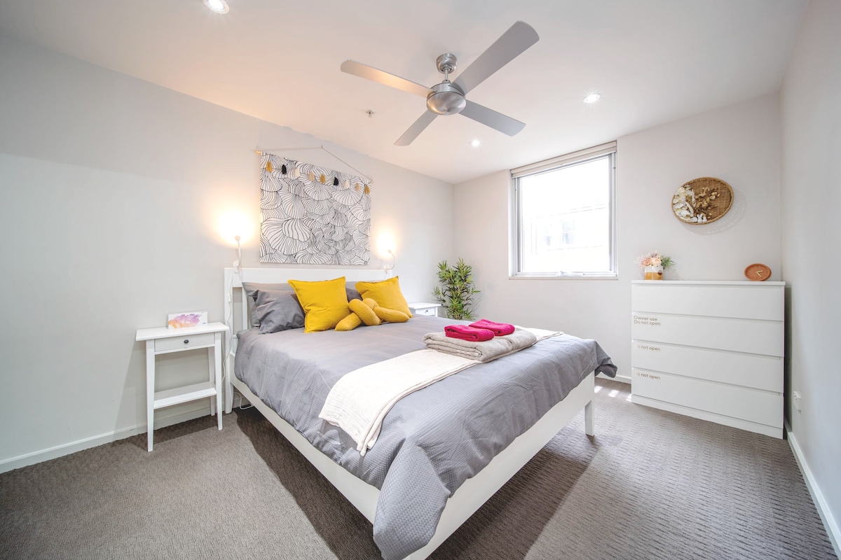 The bedroom features a queen-size bed adorned with yellow throw pillows and a cozy blanket. A bedside table and a dresser are positioned nearby. Natural light enters through a large window, and a plant adds a touch of greenery to the space. A ceiling fan enhances airflow.